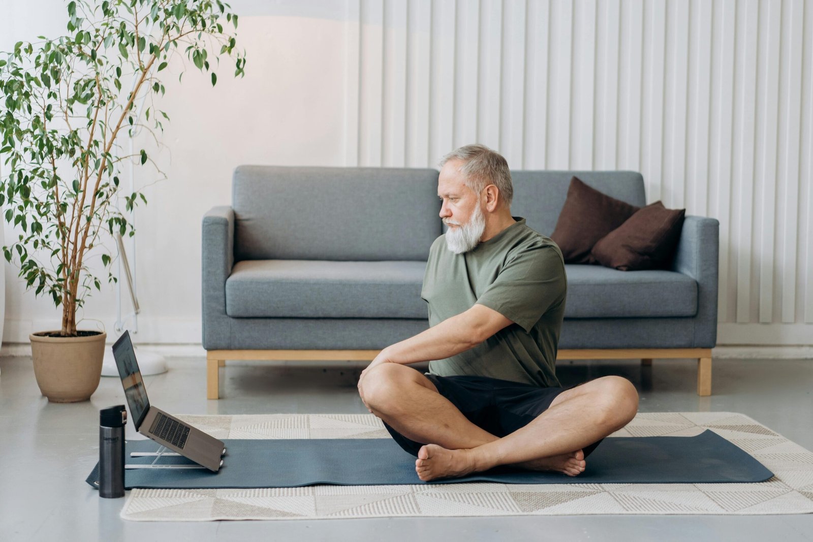 Senior man doing yoga on a mat at home, facing a laptop for virtual guidance.