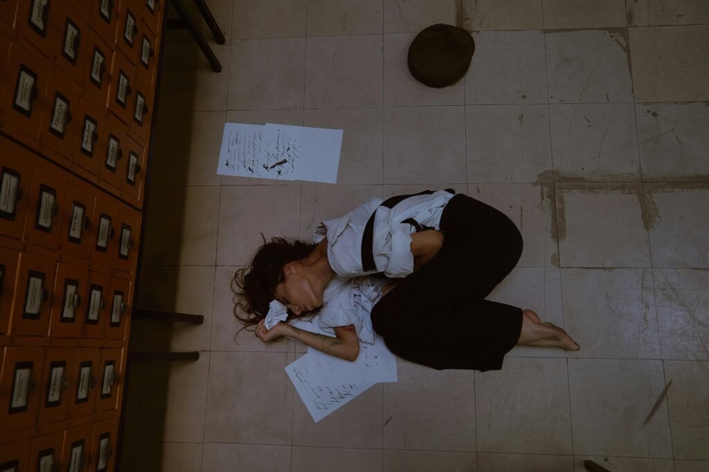 A young woman lies on the floor surrounded by papers, depicting themes of stress and exhaustion.